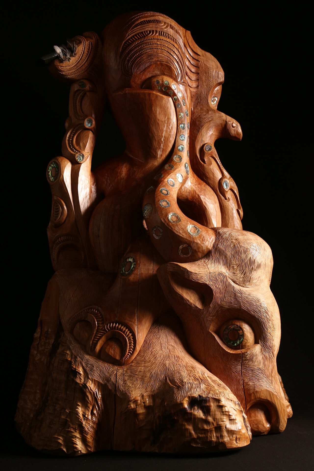Whakairo by Kade Cowper, carved from a single trunk. A rider, masked below the eyes, holds a bull by both horns: one horn becomes an octopus tentacle inlaid with pāua that enters the rider, the other becomes a manu clasped in three fingers, its wing becoming the moko on the rider's forehead. The rider's other hand holds a toki, and the bull bows down onto its hooves.