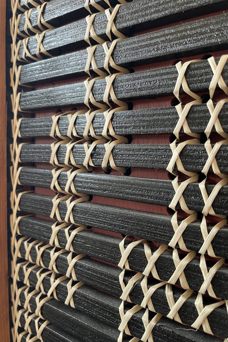 Tukutuku by Aka Whiro: black slats bound with cream kiekie in crossed stitches, seen at a low angle over an ochre red board.