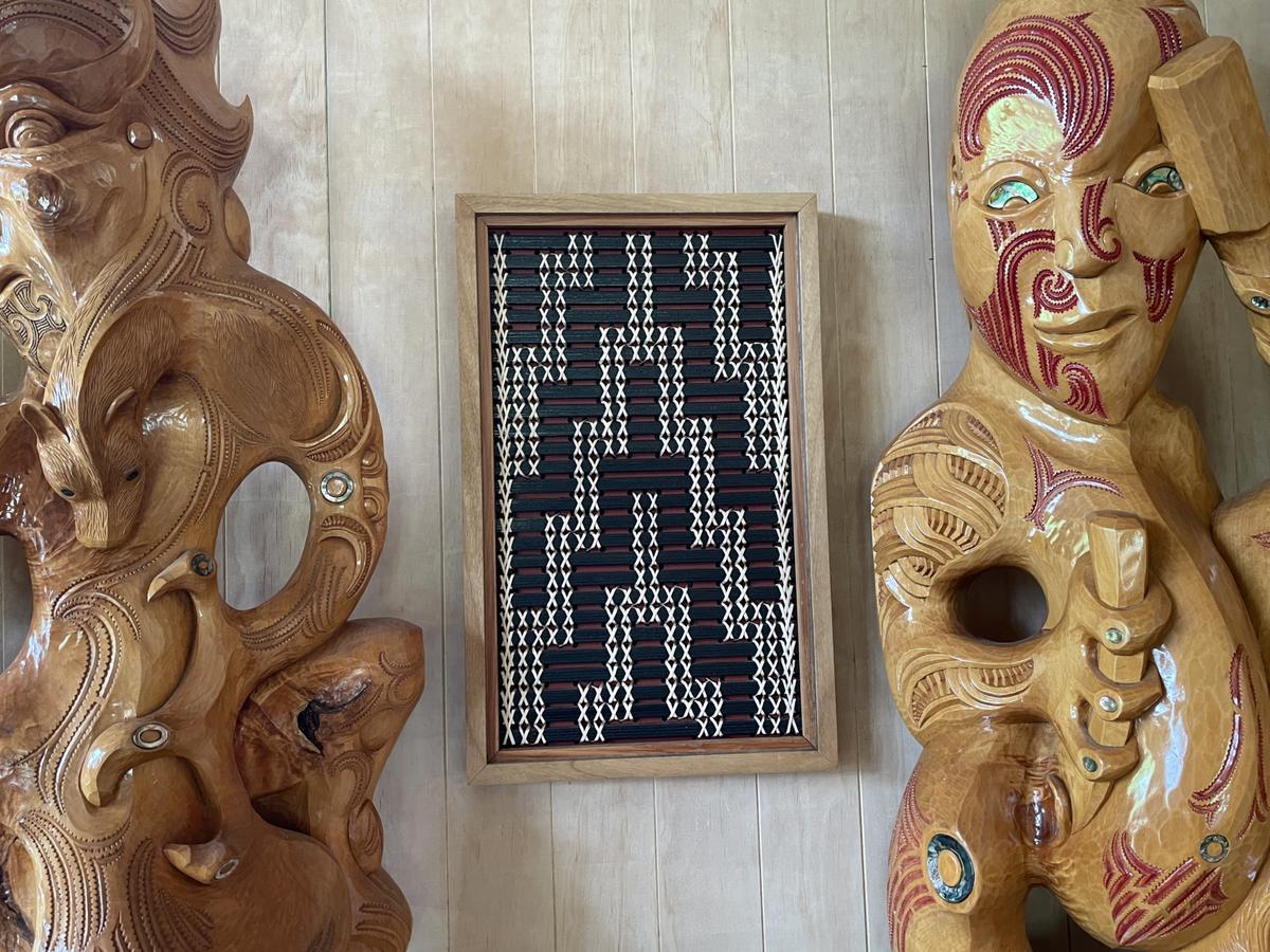 Poutama by Aka Whiro, hanging on a timber wall between two large carved figures.