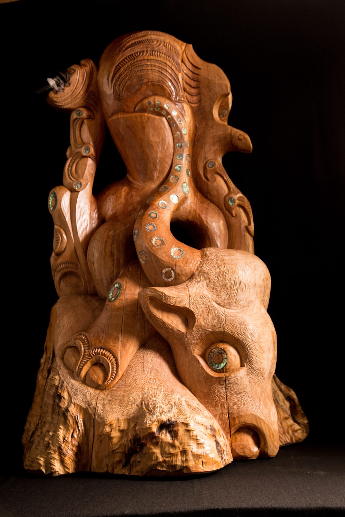 Te Ara Whakaiti, three-quarter view: the rider grips the bull by both horns, the pāua-set tentacle curving down to the bull's head, the tōtara left raw below.