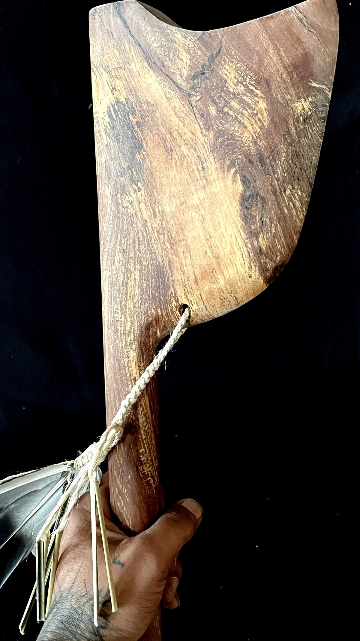 Tewhatewha by Kade Cowper, held upright in a hand: a figured timber head, with a braided cord and feather tuft.